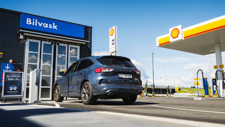 A blue hatchback-type car shown from the back corner in front of a car wash entry at a Shell petrol station. An A-stand with text in Norwegian is visible on the left side of the car. A petrol prices sign with a Shell logo is visible on the background. A part of a pentice with a Shell logo of a refuelling station is visible in the top right corner of the photo.