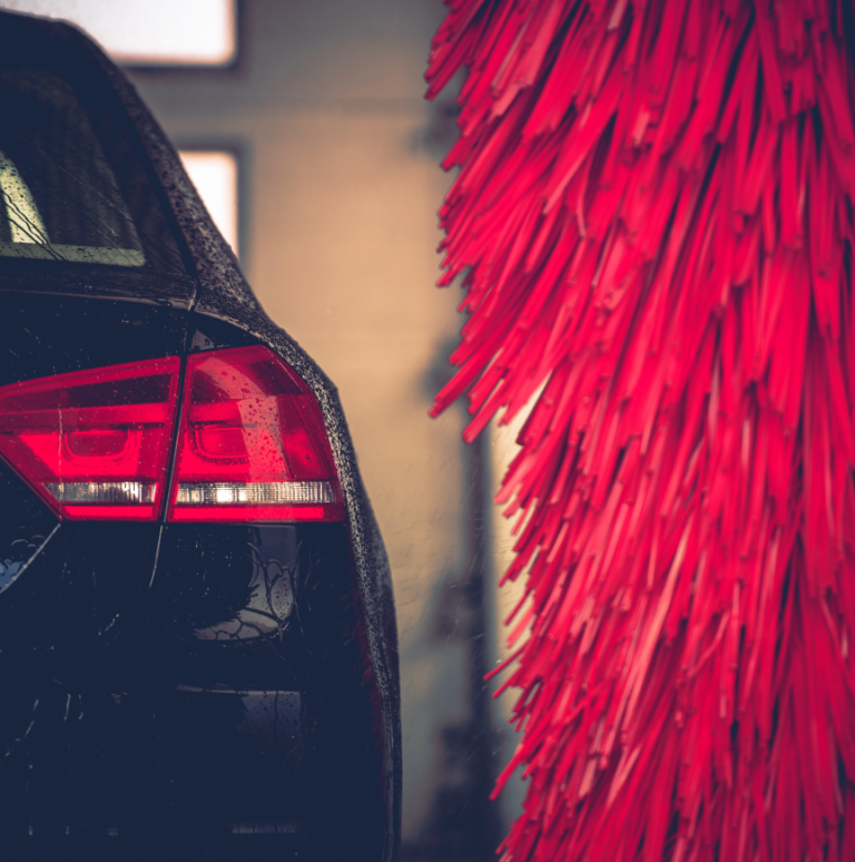 The right rear corner of a black car. One rear light, a part of the rear window, and a part of the trunk door are visible. A part of a red cylindrical car wash brush is shown on the right side of the car.