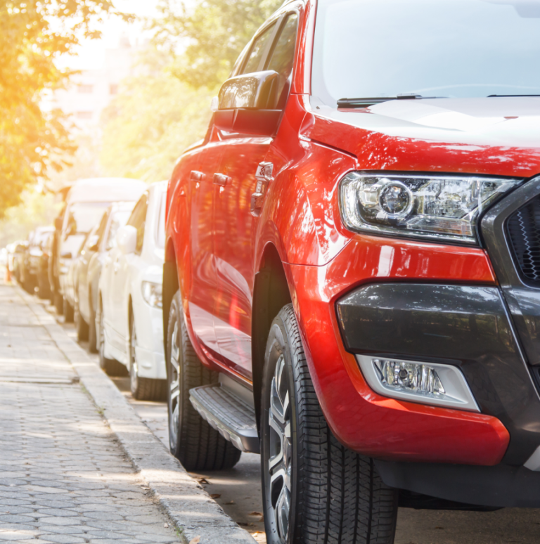 Cars parked on the side of a sidewalk. On the front first in the row, a red car is shown from the front corner. The side and a part of the mask are visible.